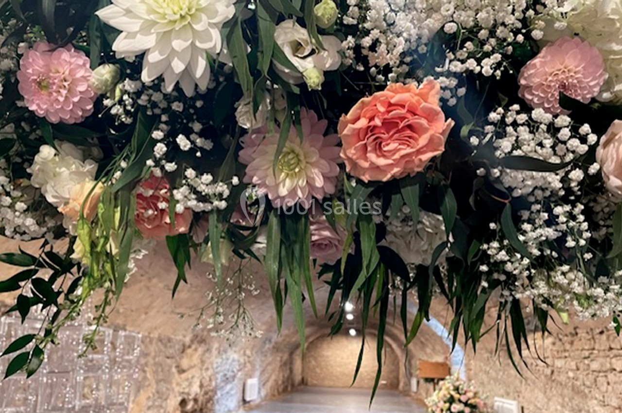 Arrangement floral suspendu avec roses, dahlias et gypsophiles, dans une salle voûtée en pierre éclairée.