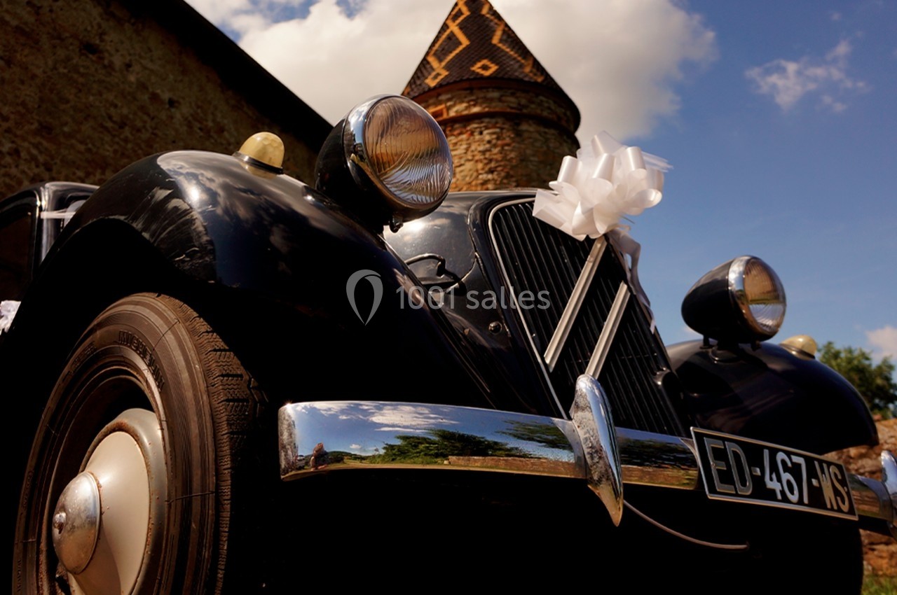 Voiture ancienne noire décorée pour un mariage, stationnée devant une tour en pierre sous un ciel bleu.