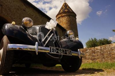 Scooter rose décoré de fleurs roses et blanches, stationné près d'un mur en pierre et d'une porte en bois.