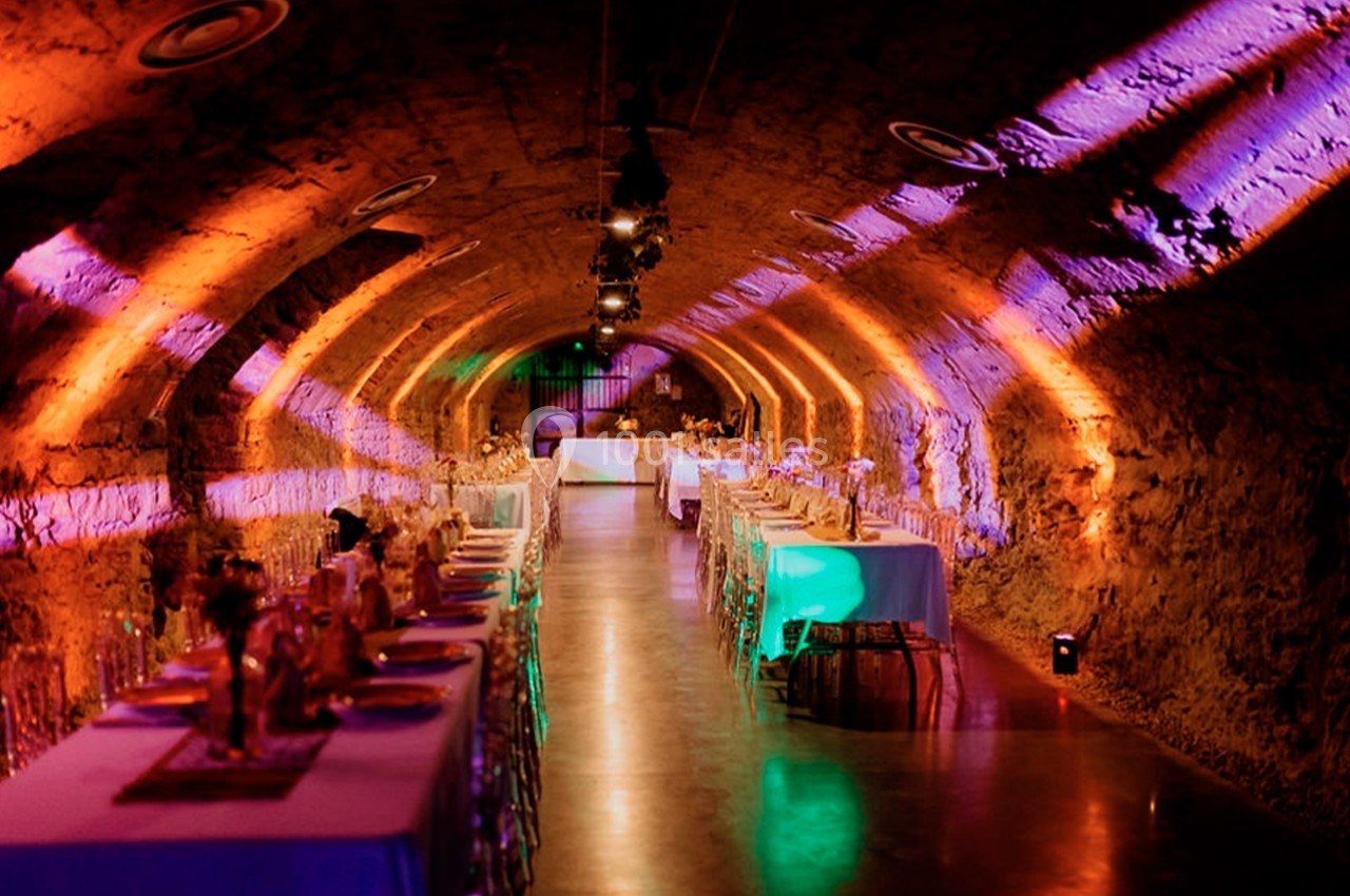 Salle voûtée en pierre éclairée par des lumières colorées, aménagée avec des tables dressées pour un événement.