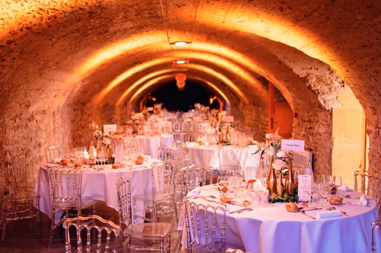 Salle voûtée décorée pour un événement, avec tables rondes dressées, chaises transparentes et éclairage chaleureux.