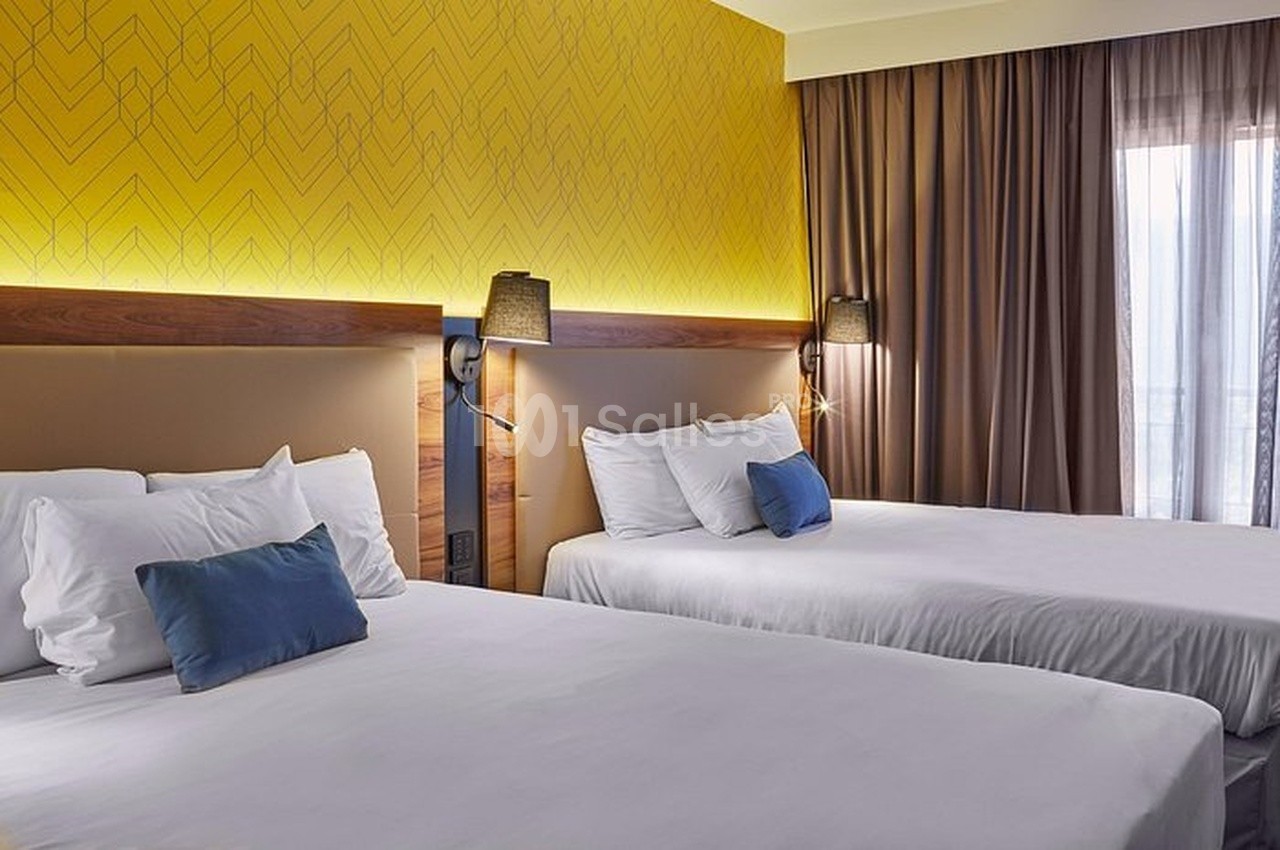 Deux lits doubles avec draps blancs, coussins bleus et mur jaune décoratif, éclairés par des lampes murales.