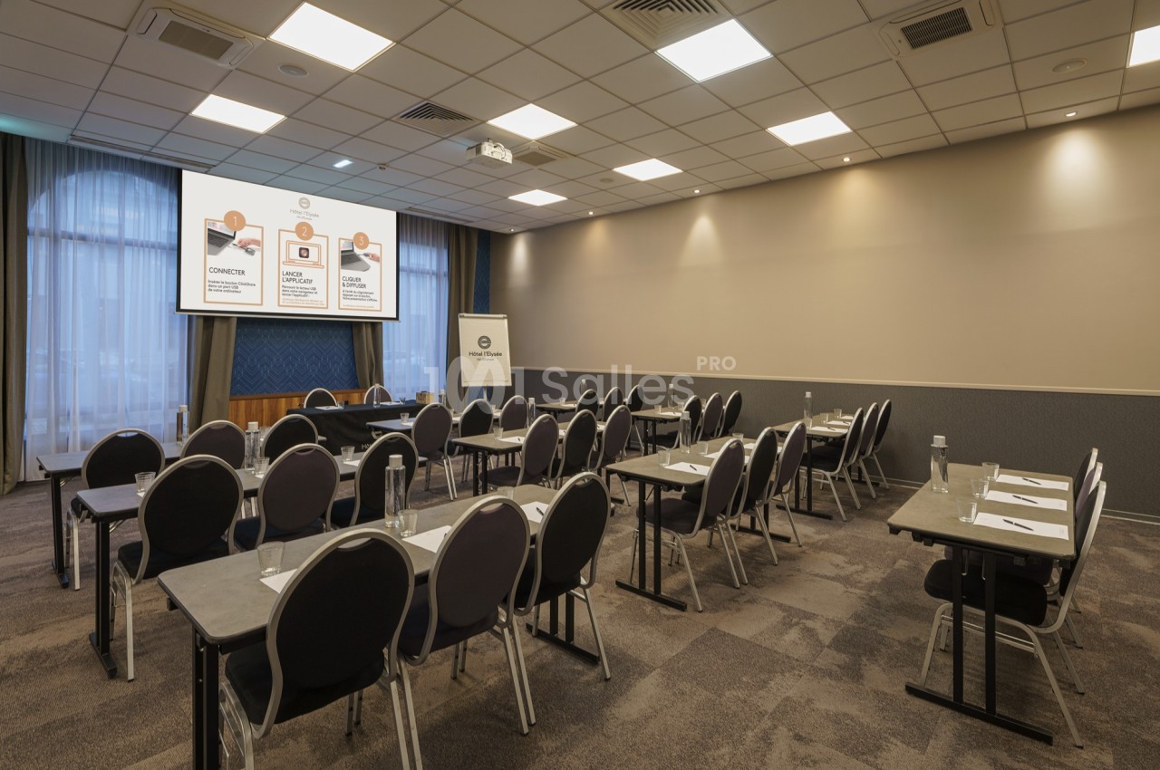 Salle de conférence équipée de tables, chaises, écran de projection et éclairage artificiel.