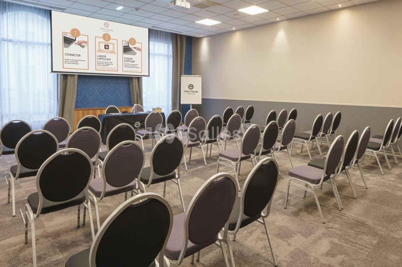 Salle de conférence avec des rangées de chaises vides, un écran de projection et un pupitre à l'avant.