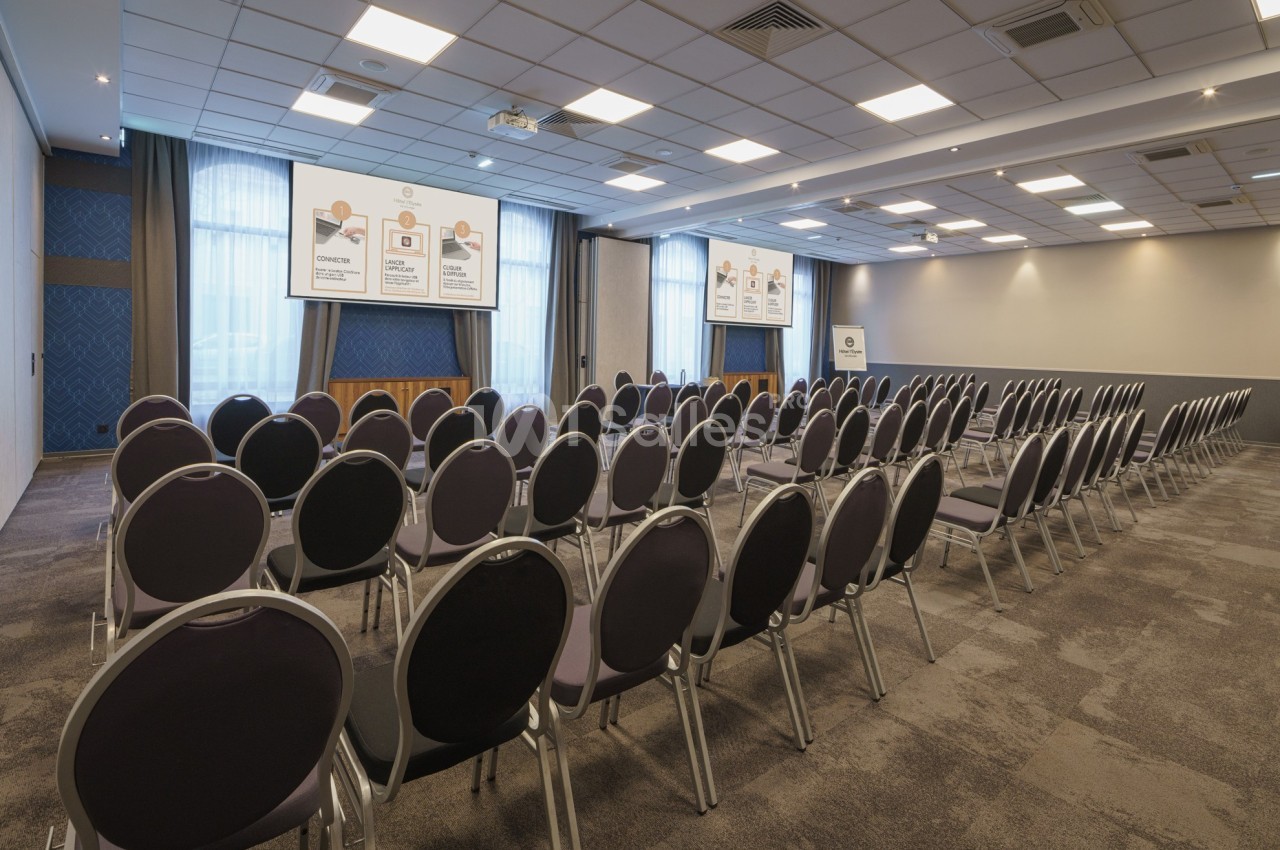 Salle de conférence vide avec des rangées de chaises alignées face à trois écrans de projection allumés.