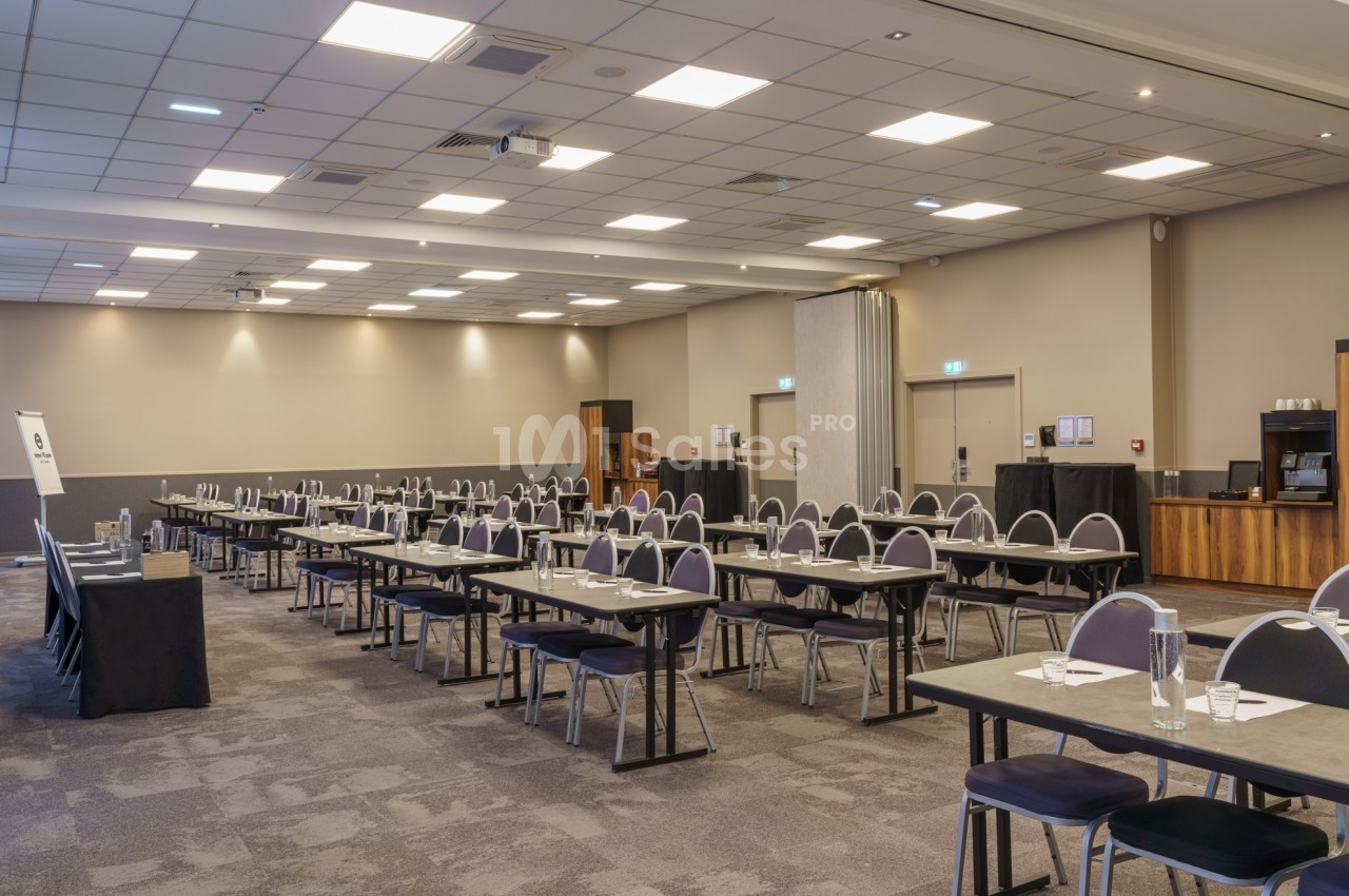 Salle de conférence vide avec des tables et des chaises alignées, éclairée par des plafonniers.