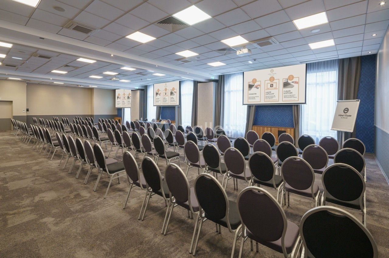 Salle de conférence vide avec des rangées de chaises noires, un écran de projection et des affiches explicatives au mur.