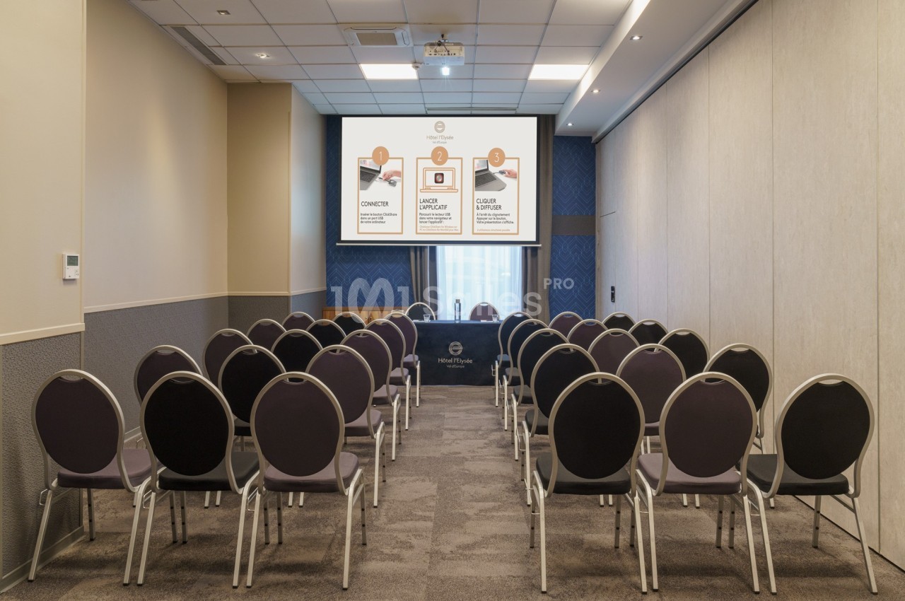 Salle de conférence vide avec des rangées de chaises alignées face à un écran de projection allumé.