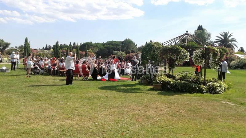 Cérémonie en plein air avec des invités assis, un officiant debout et une pergola décorée de fleurs au premier plan.