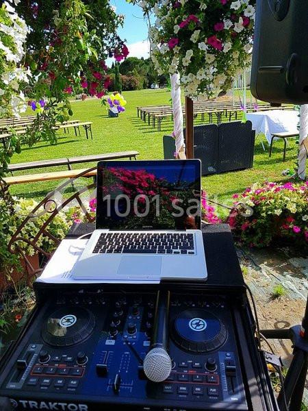 Poste de DJ en extérieur avec ordinateur portable, table de mixage et micro, entouré de fleurs et bancs sur pelouse.