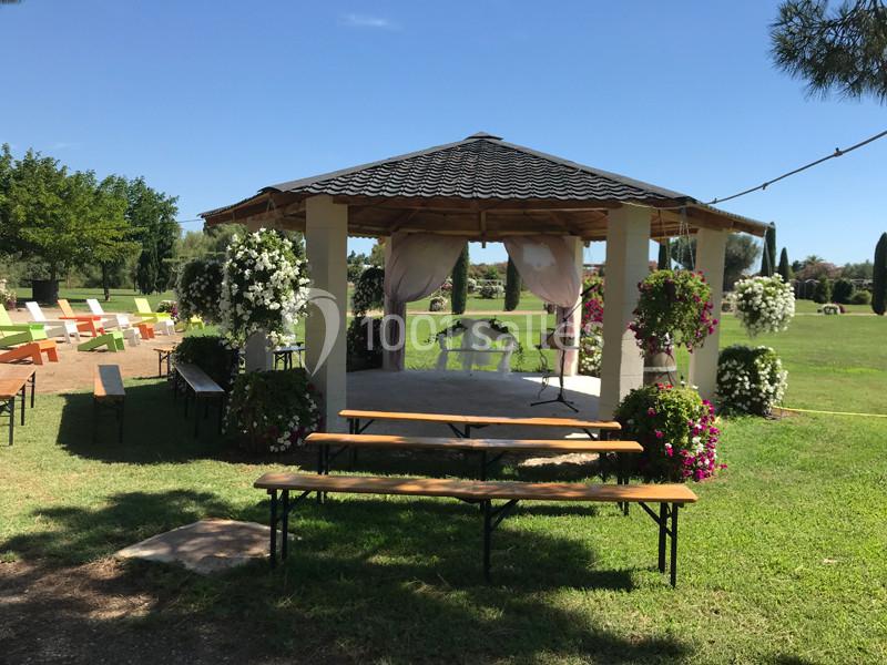 Kiosque en bois décoré de fleurs, entouré de bancs et de chaises, situé dans un jardin verdoyant par temps ensoleillé.