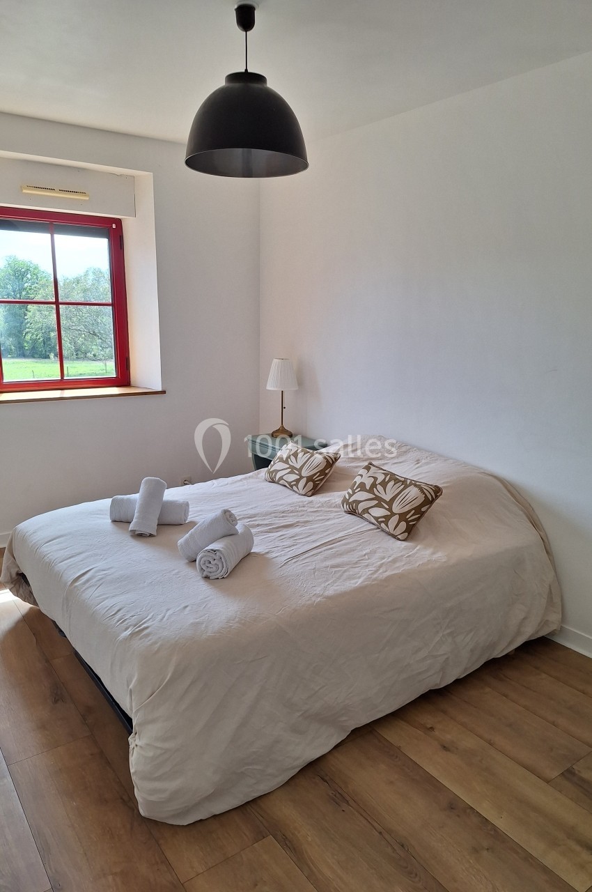 Chambre lumineuse avec un lit double, draps beiges, coussins décoratifs, lampe de chevet et fenêtre donnant sur un paysage…