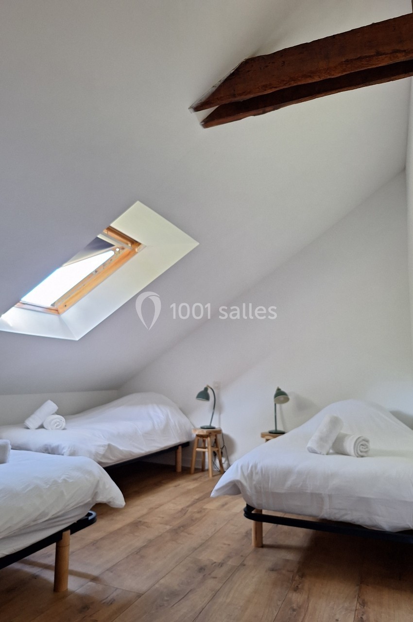 Chambre mansardée avec trois lits simples, parquet en bois, poutre apparente et fenêtre de toit laissant entrer la lumière.
