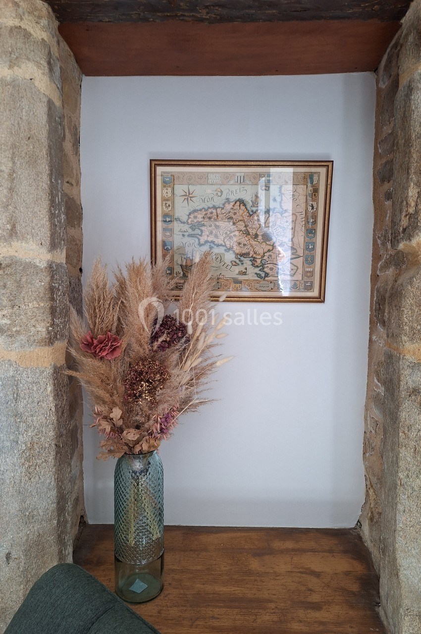 Vase en verre vert contenant un bouquet de fleurs séchées, posé sur une étagère entre deux murs en pierre, avec une carte…