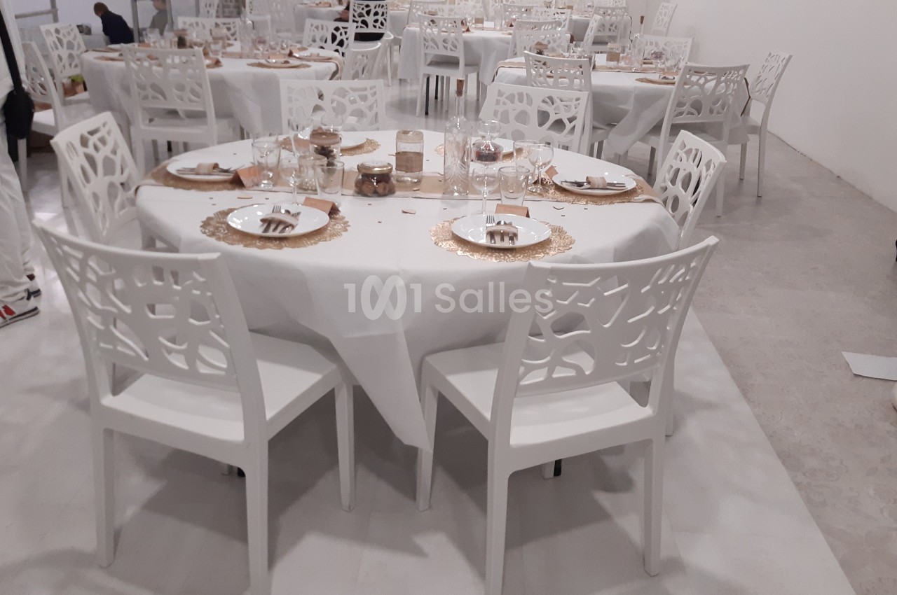 Salle de réception avec tables rondes dressées, nappes blanches, chaises blanches ajourées et décoration dorée.