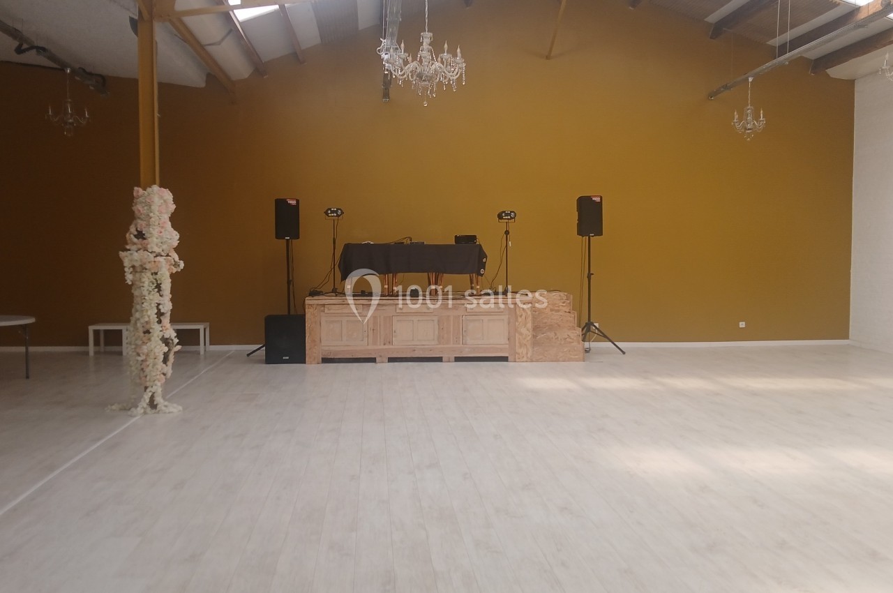 Salle spacieuse avec parquet clair, murs jaunes, plafond industriel, et une scène équipée de matériel audio.