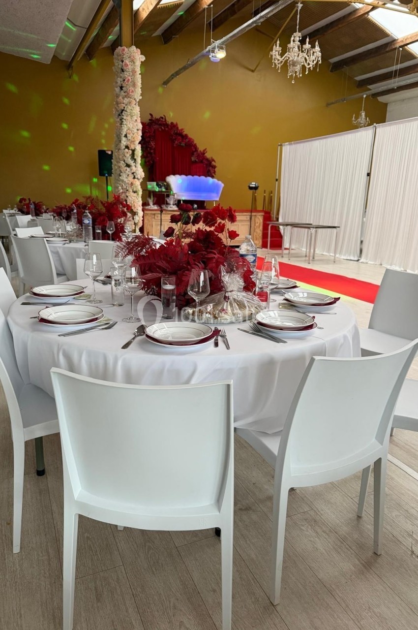 Salle de réception décorée avec des tables rondes dressées, chaises blanches, fleurs rouges et rideaux blancs en arrière…
