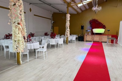 Salle de réception décorée avec des tables blanches, des colonnes fleuries et un tapis rouge menant à une estrade.