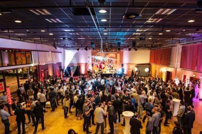 Salle de réception avec tables rondes dressées, éclairage coloré et scène équipée pour un événement.