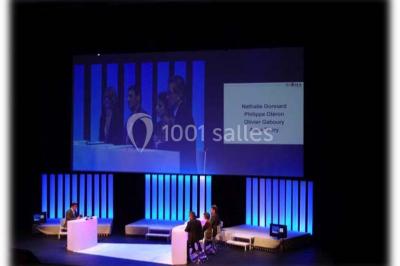 Salle de réception avec tables rondes dressées, éclairage coloré et scène équipée pour un événement.