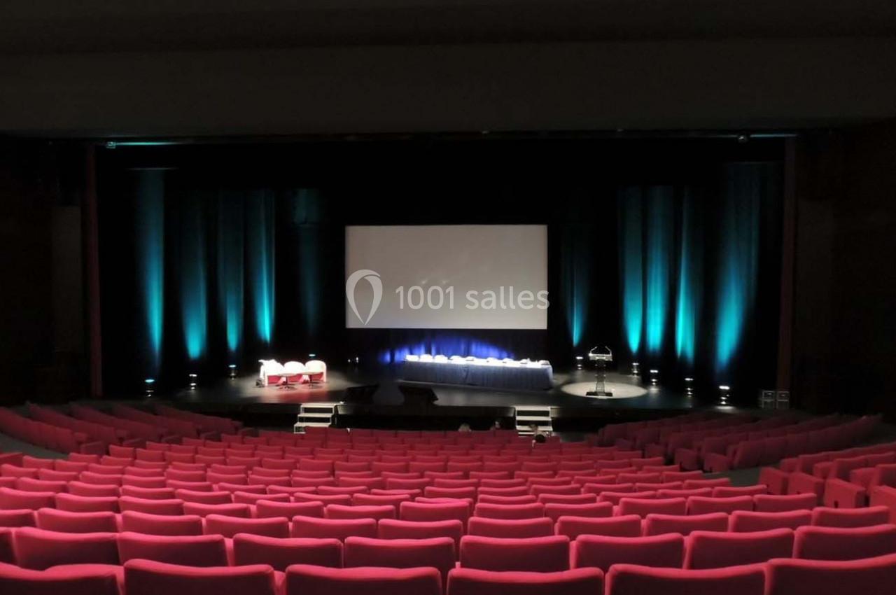 Salle de théâtre vide avec sièges rouges, scène éclairée en bleu, écran blanc et pupitre sur le côté.