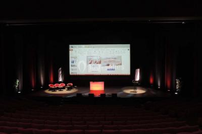 Salle de réception avec tables rondes dressées, éclairage coloré et scène équipée pour un événement.