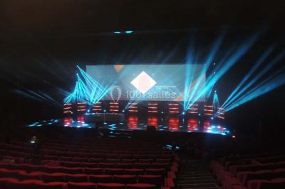 Salle de réception avec tables rondes dressées, éclairage coloré et scène équipée pour un événement.