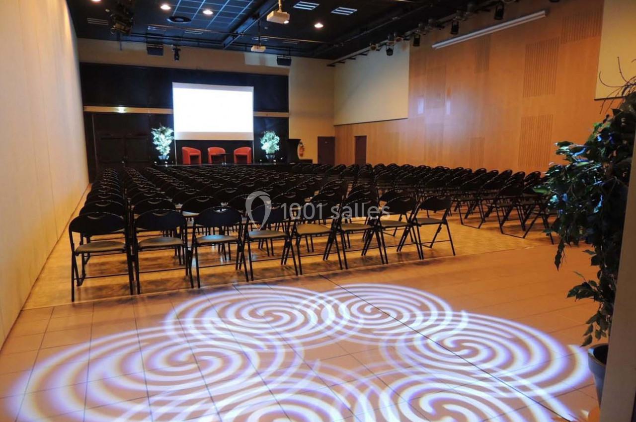 Salle de conférence avec rangées de chaises noires, écran de projection allumé et éclairage décoratif au sol.