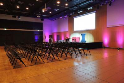 Salle de réception avec tables rondes dressées, éclairage coloré et scène équipée pour un événement.