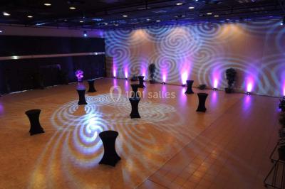 Salle de réception avec tables rondes dressées, éclairage coloré et scène équipée pour un événement.