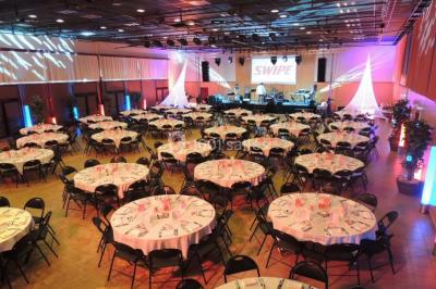 Salle de réception avec tables rondes dressées, éclairage coloré et scène équipée pour un événement.