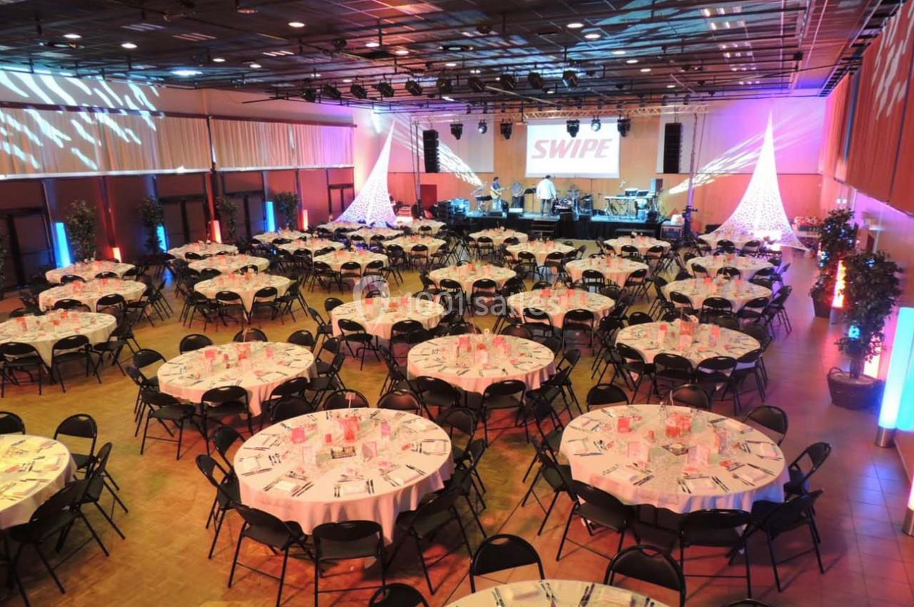 Salle de réception avec tables rondes dressées, éclairage coloré et scène équipée pour un événement.