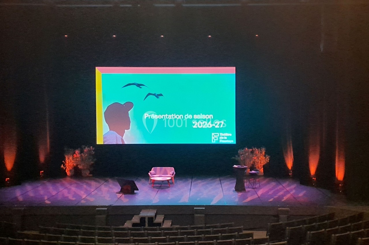 Scène de théâtre éclairée avec un écran affichant ’Présentation de saison 2026-2027’, fauteuil et pupitre au centre.
