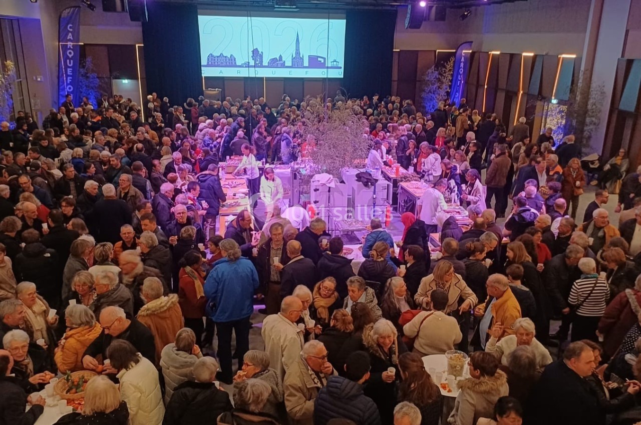 Foule rassemblée dans une grande salle éclairée, avec un buffet central et un écran projetant une image en arrière-plan.