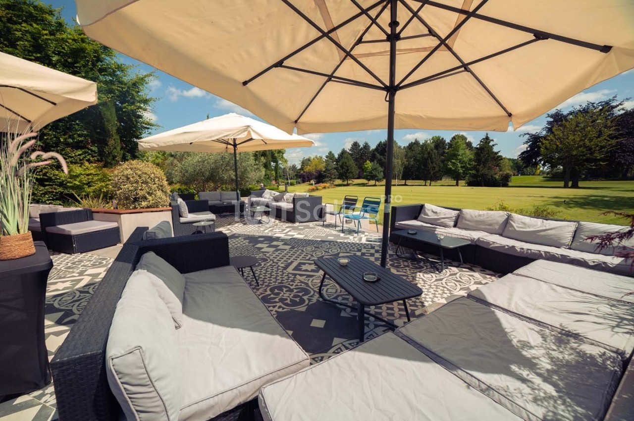 Terrasse aménagée avec canapés, tables basses et parasols, donnant sur un grand jardin verdoyant par temps ensoleillé.