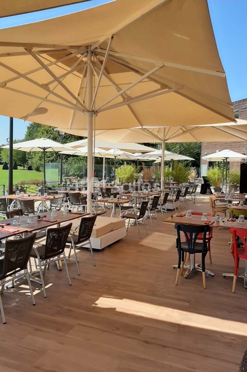 Terrasse d'un restaurant avec tables dressées, chaises en osier et grands parasols, entourée de verdure par temps ensoleillé.