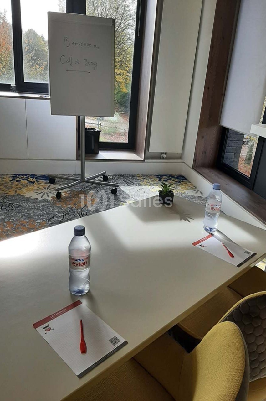 Salle de réunion lumineuse avec table, chaises, bouteilles d'eau, blocs-notes, stylos et tableau blanc près d'une fenêtre.