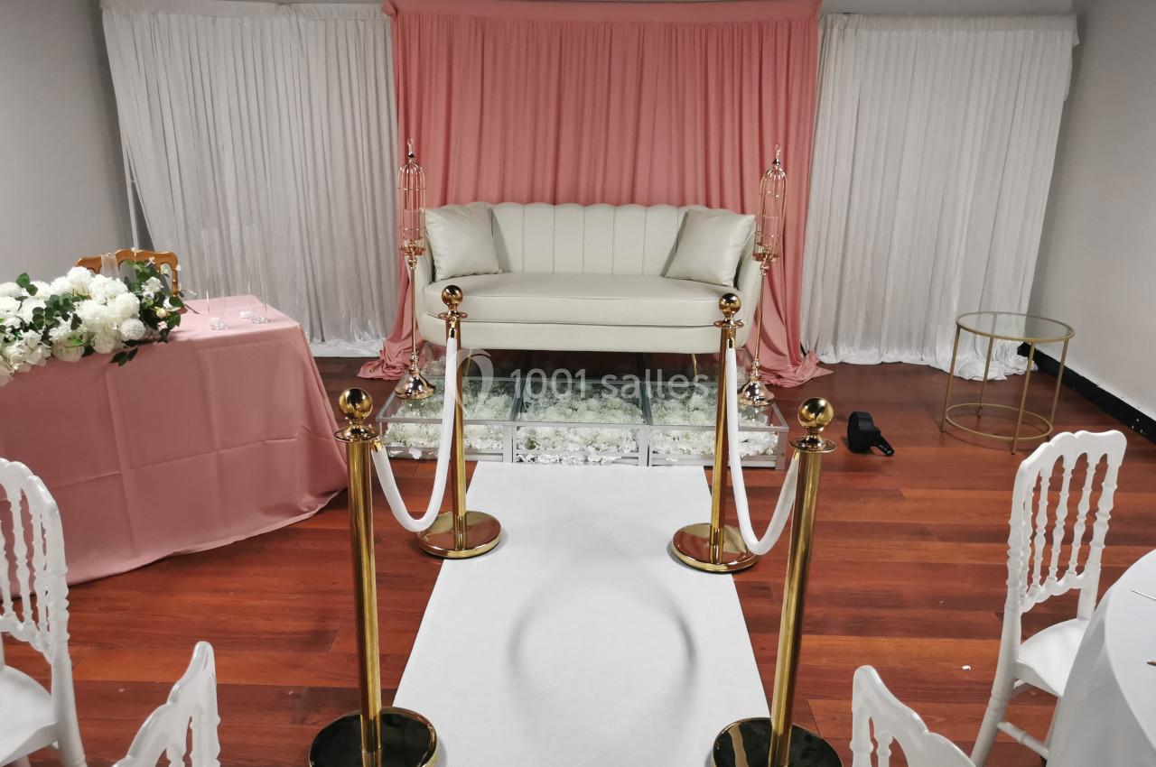 Canapé blanc devant un rideau rose et blanc, encadré de poteaux dorés avec cordons, sur un tapis blanc.