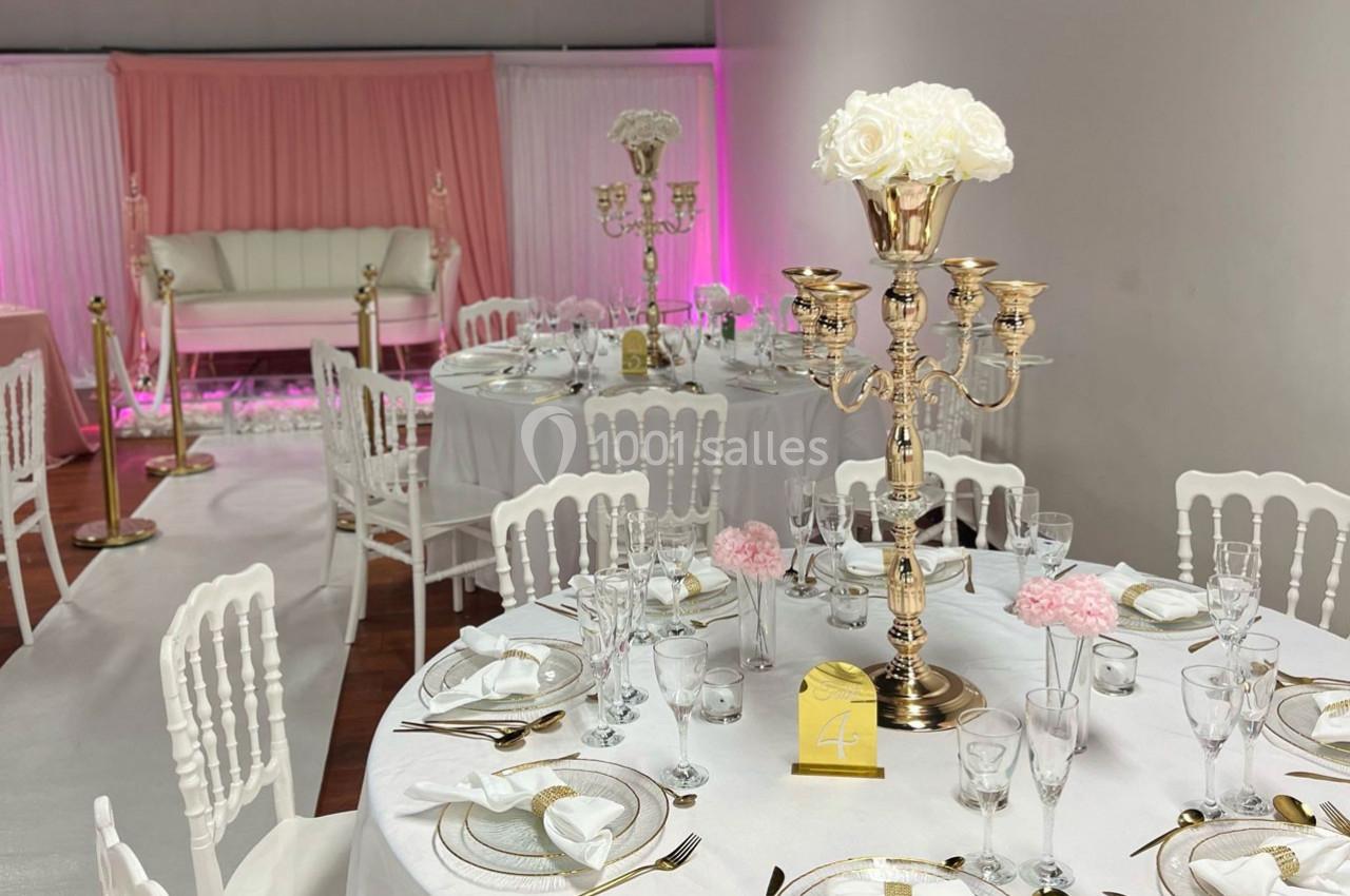 Salle de réception décorée avec des tables rondes, nappes blanches, chandeliers dorés et un fond rose élégant.