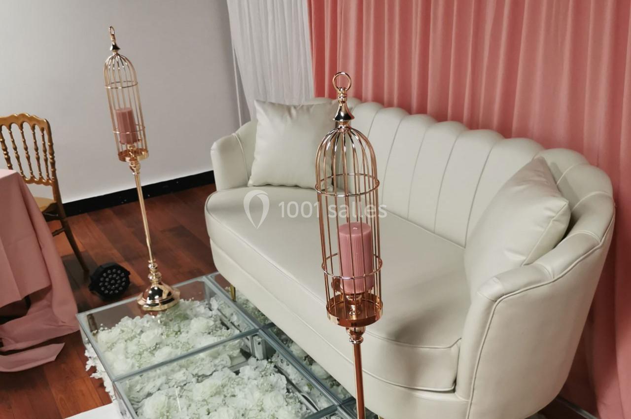 Canapé blanc devant un rideau rose, entouré de bougeoirs dorés en forme de cages et d'une table en verre.