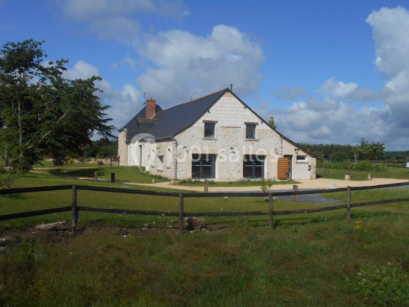 Maison en pierre avec toit en ardoise, entourée de pelouse, clôture en bois et ciel partiellement nuageux.