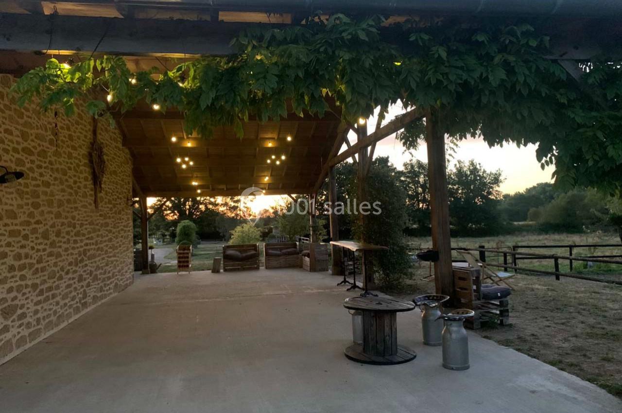Terrasse couverte en bois avec guirlandes lumineuses, entourée de verdure et donnant sur un paysage champêtre au coucher du…