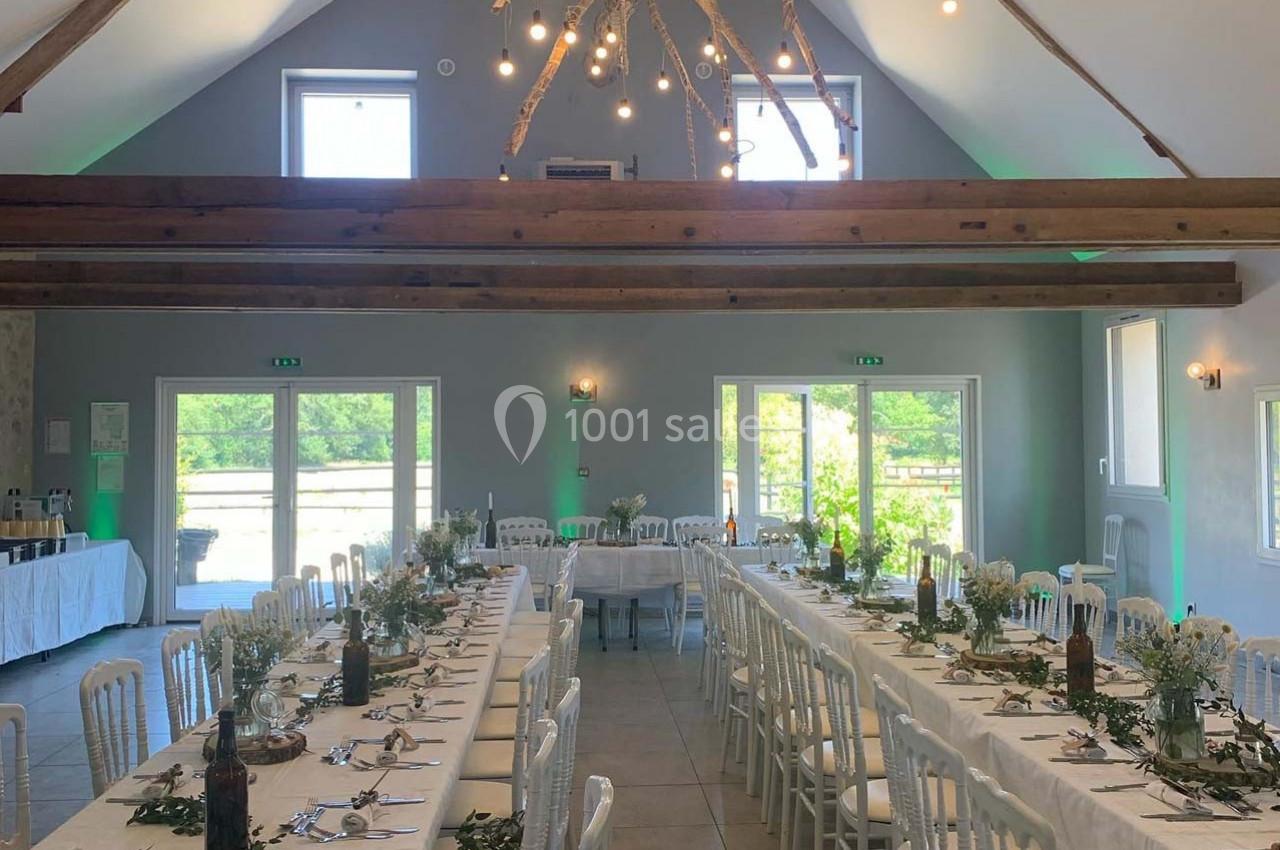 Salle de réception lumineuse avec de longues tables décorées de fleurs et de verdure, sous un plafond en poutres apparentes.