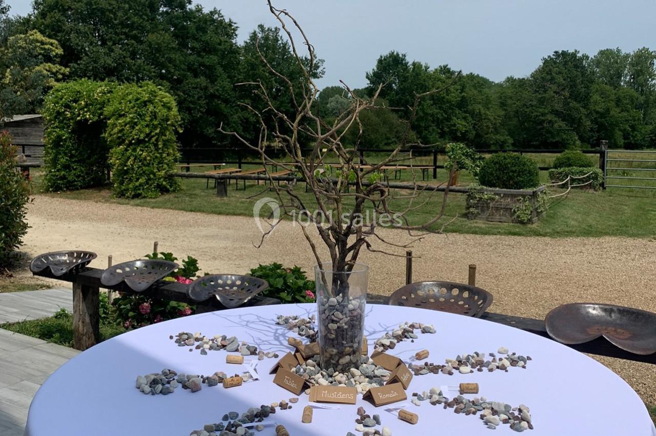 Centre de table avec branches sèches dans un vase, entouré de pierres et bouchons, sur une table ronde en extérieur.
