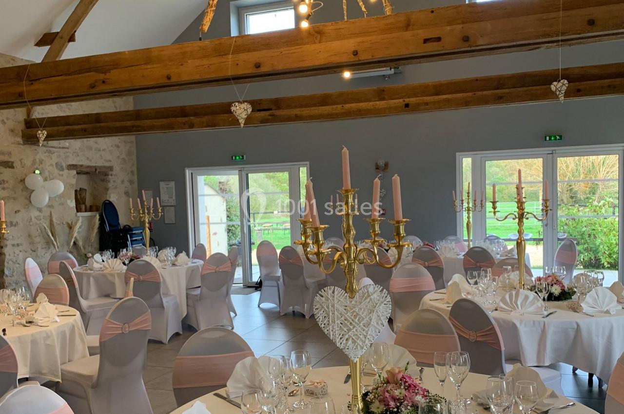 Salle de réception décorée avec des tables rondes, nappes blanches, chaises ornées de rubans roses et chandeliers dorés.