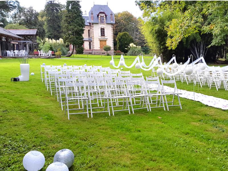 Chaises blanches alignées en extérieur pour une cérémonie, avec une allée décorée et un bâtiment en arrière-plan.