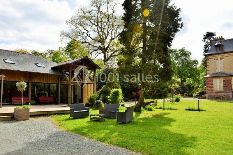 Espace extérieur avec salon de jardin, pelouse, arbres et bâtiments en arrière-plan sous un ciel partiellement nuageux.