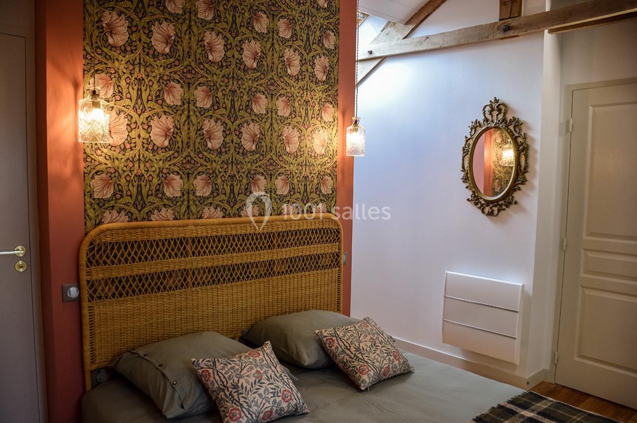 Chambre avec lit double, tête de lit en rotin, papier peint floral, miroir ovale et poutres apparentes.