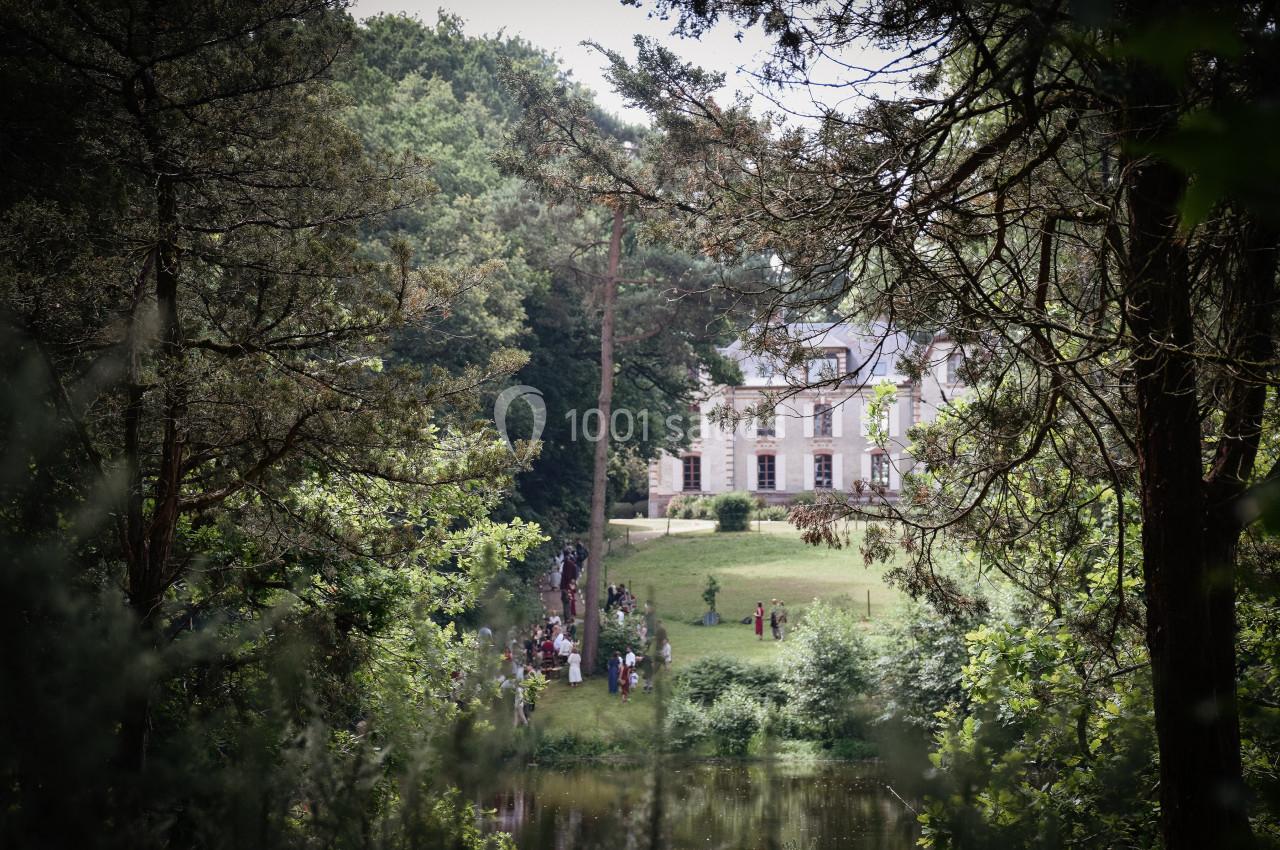 Manoir entouré d'arbres, avec des personnes rassemblées sur une pelouse près d'un étang.