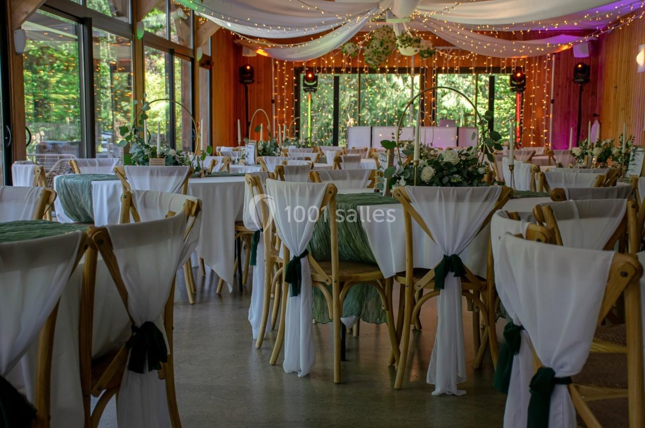 Salle de réception décorée avec des tables rondes, nappes blanches, chaises en bois et guirlandes lumineuses suspendues.